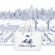Load image into Gallery viewer, Tote bag with 'Côte d'Aqua' branding in front of a cityscape illustration.